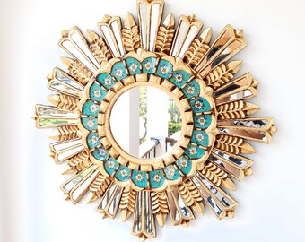May include: A gold sunburst mirror with a turquoise and white floral design around the mirror. The mirror is surrounded by a gold frame with a sunburst design.