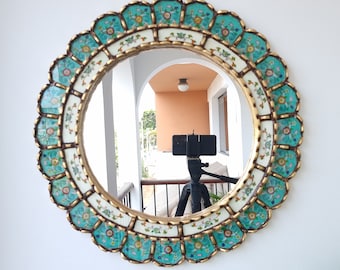 May include: Round decorative mirror with a scalloped edge. The frame features turquoise, white, and gold accents with floral patterns. The mirror is hanging on a white wall.