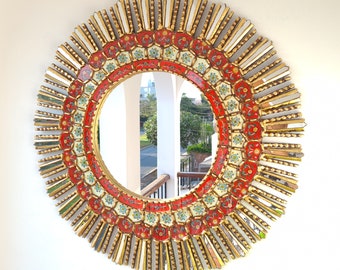 May include: A round, gold-framed mirror with a red, white, and blue mosaic design. The mirror is surrounded by a ring of gold-painted, pointed pieces of glass.