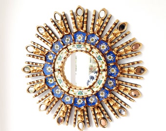 May include: A gold sunburst mirror with blue and white floral tile accents. The mirror has a round shape and is surrounded by a decorative frame.