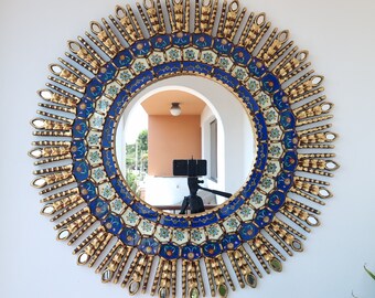 May include: A round decorative mirror with a sunburst design. The mirror has a gold-colored frame with blue and white accents. The center mirror reflects a view of a balcony and a tripod.