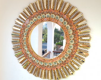May include: A round decorative mirror with a sunburst design. The mirror has a central circular reflective surface surrounded by a detailed frame. The frame features a repeating pattern of gold, white, and coral-colored elements, creating a striking visual effect.