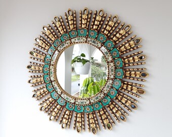 May include: A gold sunburst mirror with a round, teal and white mosaic tile design. The mirror reflects a potted plant and a view of a house.