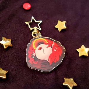 Puede incluir: Un llavero dorado con un cierre en forma de estrella y un colgante con un personaje pelirrojo con un halo. El colgante está rodeado de estrellas doradas y un botón rojo sobre un fondo morado oscuro.