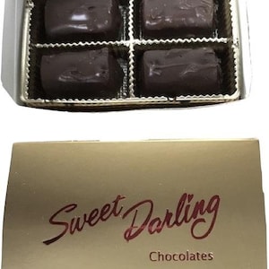 Puede incluir: Una caja dorada con las palabras "Sweet Darling Chocolates" impresas en la parte delantera. La caja contiene cuatro cuadrados de chocolate negro.