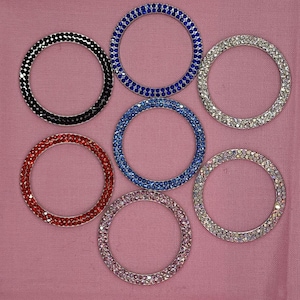 May include: Seven rhinestone-encrusted rings in various colors, including black, blue, red, and silver. The rings are arranged in a circle on a pink surface.