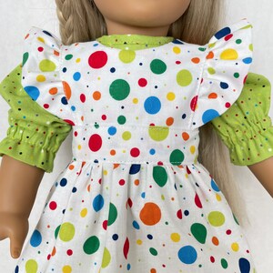 May include: A white dress with a green polka dot pattern. The dress has a ruffled neckline and a green sash. The dress is being worn by a doll.