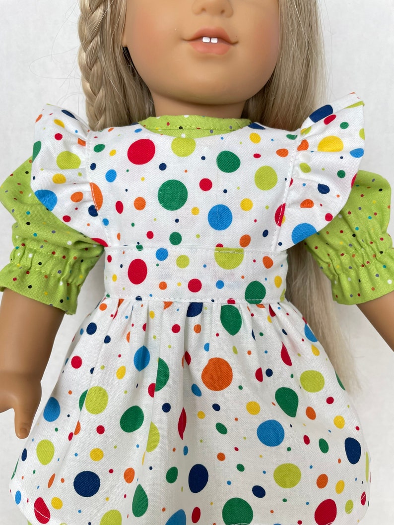 May include: A white dress with a green polka dot pattern. The dress has a ruffled neckline and a green sash. The dress is being worn by a doll.