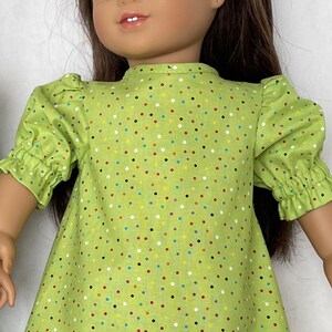 May include: A green dress with colourful polka dots for a doll. The dress has puffed sleeves and a round neckline.