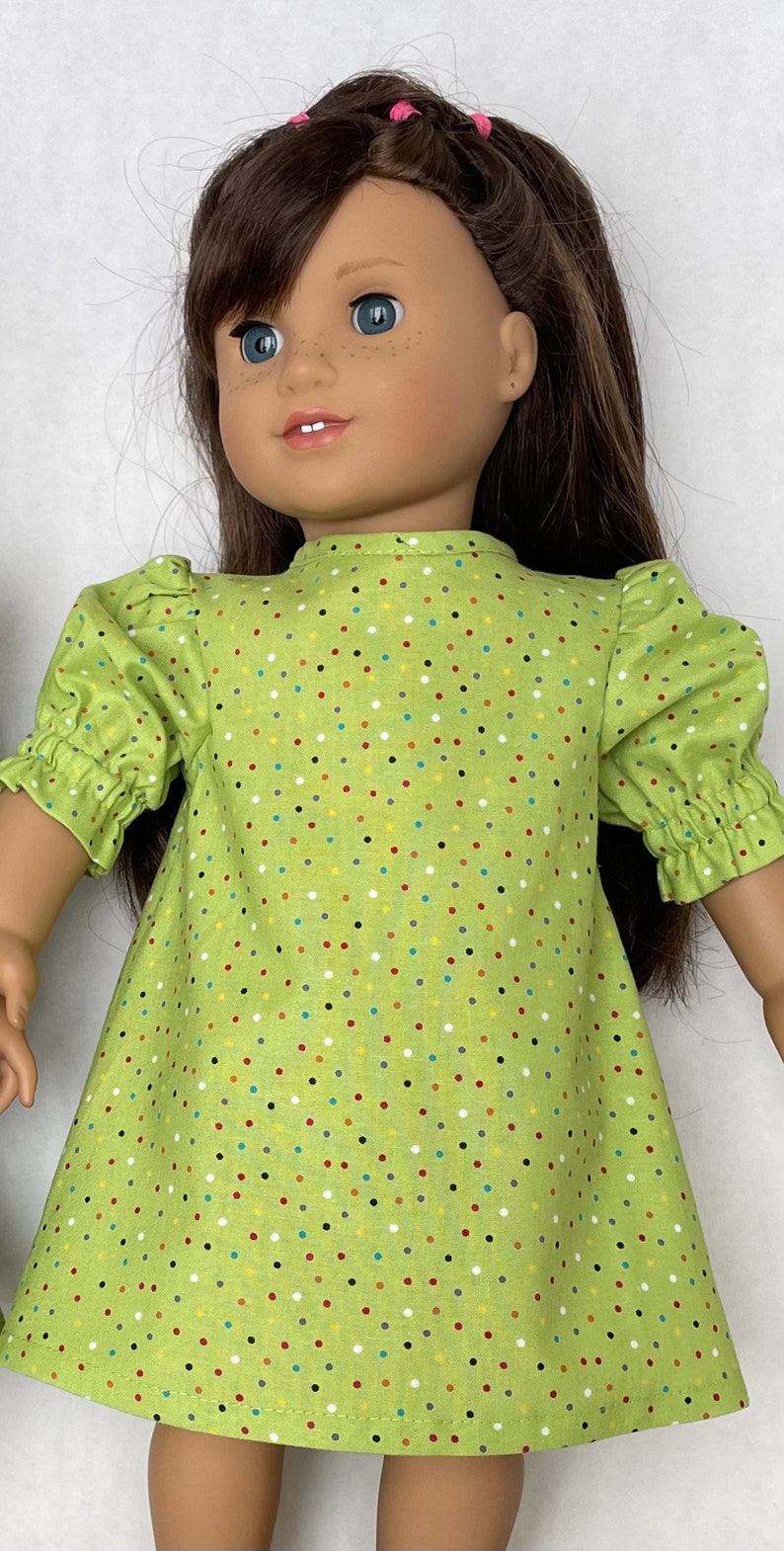 May include: A green dress with colourful polka dots for a doll. The dress has puffed sleeves and a round neckline.