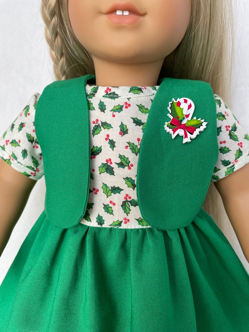 May include: A green dress with a white holly print top and a green vest. The dress has a candy cane and holly pin.