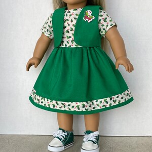 May include: A green dress with a white and red holly pattern, a green vest, and a red and white holly pin. The doll is wearing green sneakers.