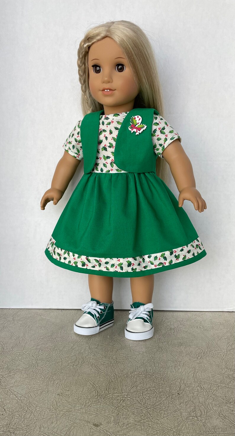 May include: A green dress with a white and red holly pattern, a green vest, and a red and white holly pin. The doll is wearing green sneakers.
