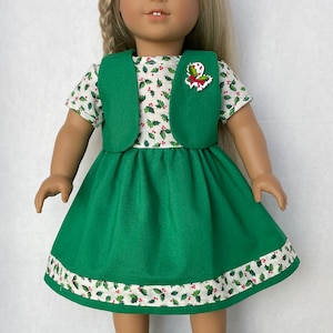May include: A green dress with a white holly pattern and a green vest. The doll is wearing white sneakers.