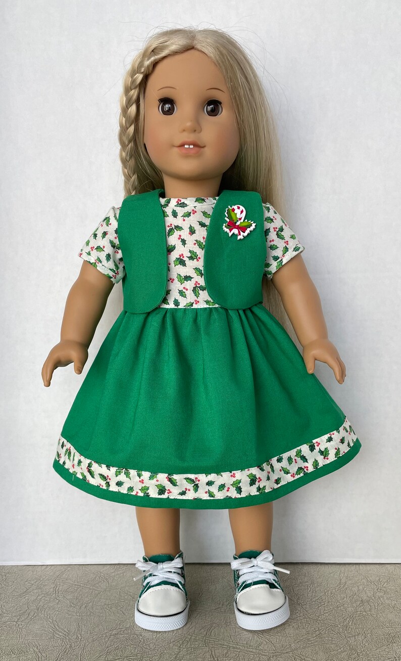 May include: A green dress with a white holly pattern and a green vest. The doll is wearing white sneakers.