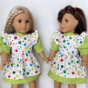 May include: Two dolls wearing white dresses with green and colourful polka dots. The dresses have ruffled sleeves and a green trim. The dolls have long brown hair and are smiling.