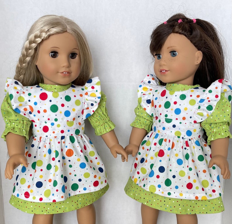 May include: Two dolls wearing white dresses with green and colourful polka dots. The dresses have ruffled sleeves and a green trim. The dolls have long brown hair and are smiling.