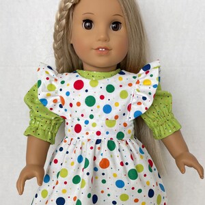 May include: A doll wearing a white dress with colourful polka dots and a green ruffled top. The dress has a green polka dot trim.