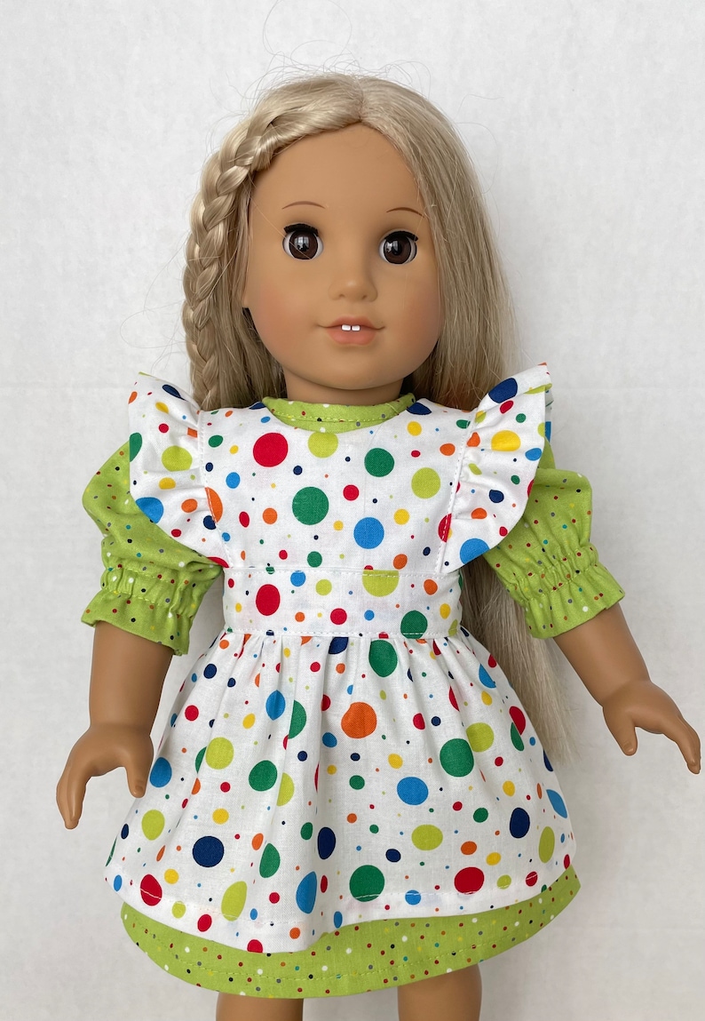 May include: A doll wearing a white dress with colourful polka dots and a green ruffled top. The dress has a green polka dot trim.