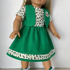 May include: A green dress with a white holly pattern and a green vest. The dress has a white trim with a holly pattern. The doll is wearing green sneakers with white laces.