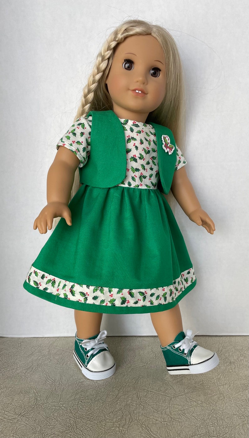 May include: A green dress with a white holly pattern and a green vest. The dress has a white trim with a holly pattern. The doll is wearing green sneakers with white laces.