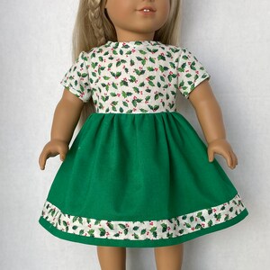 May include: A doll wearing a green and white dress with a holly pattern. The dress has a green skirt and a white top with red berries and green leaves. The doll is wearing white sneakers with green accents.