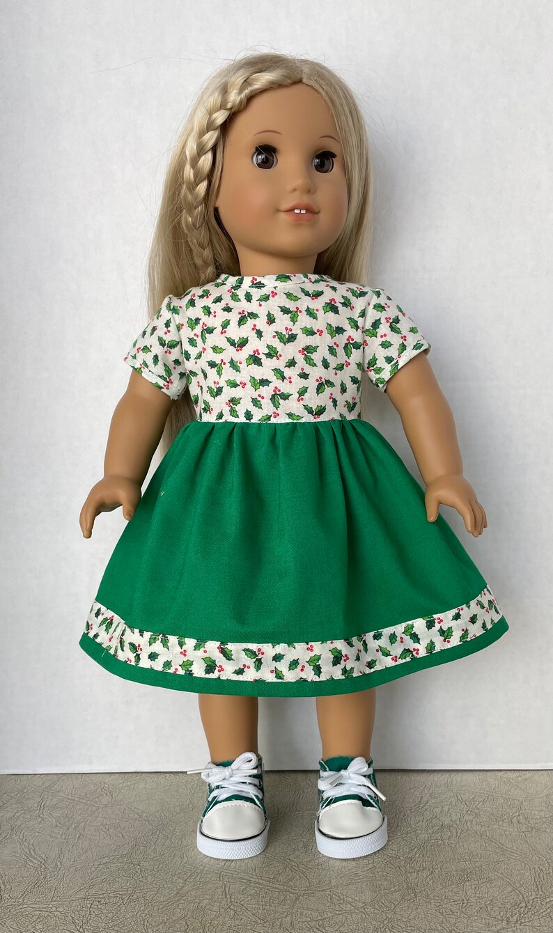 May include: A doll wearing a green and white dress with a holly pattern. The dress has a green skirt and a white top with red berries and green leaves. The doll is wearing white sneakers with green accents.