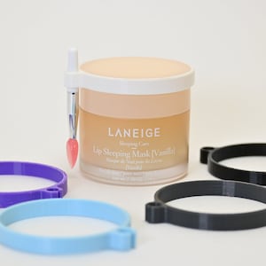 May include: A jar of Laneige Lip Sleeping Mask [Vanilla] with a lip brush applicator and three circular holders in purple, light blue, and black. The jar has a beige lid and a clear glass base. The text "Laneige" is visible on the jar.