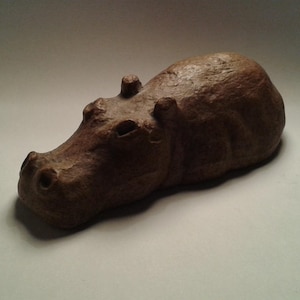 May include: A brown, textured hippo figurine. The sculpture has a detailed snout, small ears, and a rounded body. The hippo is a neutral brown color, and the surface has a rough, natural appearance. The figurine is a decorative item.