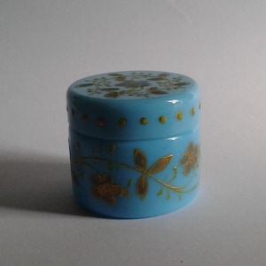 May include: A small, round, light blue jar with a lid. The jar is decorated with gold floral designs. The lid and the body of the jar have gold accents. The jar is sitting on a white surface.