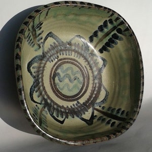 May include: A square ceramic bowl with a green glaze and black and white painted design. The design features a central circle with wavy lines and a surrounding flower-like pattern.