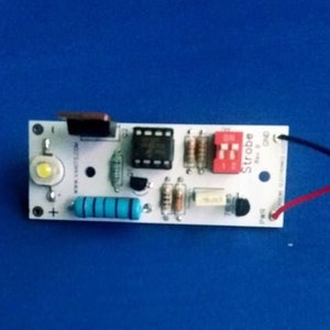 May include: A white circuit board with a red and black switch labeled "Strobe" and a blue resistor. The board has a white LED light, a black speaker, and a black transistor. The board is connected to red and black wires.