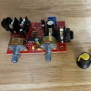 May include: A red printed circuit board for a mini guitar amplifier featuring two metallic potentiometers, input and output jacks, capacitors, and resistors. Two separate black plastic control knobs with yellow and blue inserts are included.