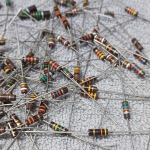 Op de afbeelding: Een grote verzameling van verschillende kleurgecodeerde weerstanden met bruine, oranje, gele, rode, groene, blauwe en zwarte banden. De weerstanden zijn willekeurig gerangschikt op een grijs oppervlak.
