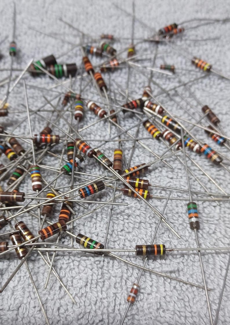 Op de afbeelding: Een grote verzameling van verschillende kleurgecodeerde weerstanden met bruine, oranje, gele, rode, groene, blauwe en zwarte banden. De weerstanden zijn willekeurig gerangschikt op een grijs oppervlak.