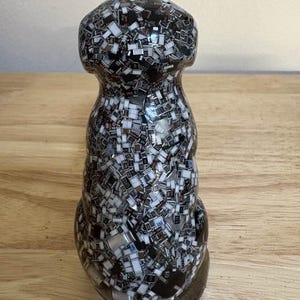 May include: A decorative, clear glass bottle stopper with a unique mosaic design. The stopper is filled with small, rectangular black, white, and silver pieces, creating a textured, abstract pattern. The base is a dark, rounded shape.