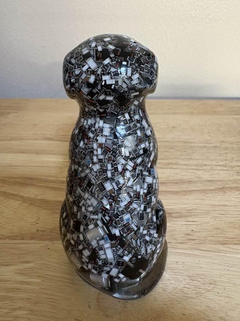 May include: A decorative, clear glass bottle stopper with a unique mosaic design. The stopper is filled with small, rectangular black, white, and silver pieces, creating a textured, abstract pattern. The base is a dark, rounded shape.