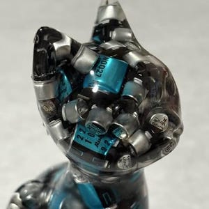 Puede incluir: Una escultura de gato de resina transparente llena de componentes electr&oacute;nicos plateados y turquesas. El gato tiene una oreja puntiaguda y un dise&ntilde;o elegante y moderno. Los componentes son visibles a trav&eacute;s de la resina transparente.