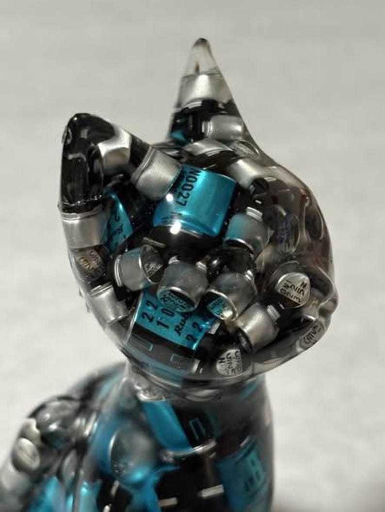 Puede incluir: Una escultura de gato de resina transparente llena de componentes electr&oacute;nicos plateados y turquesas. El gato tiene una oreja puntiaguda y un dise&ntilde;o elegante y moderno. Los componentes son visibles a trav&eacute;s de la resina transparente.