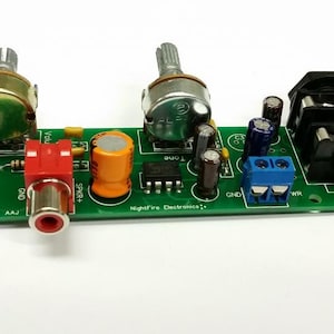 Puede incluir: Una placa de circuito verde con una toma RCA negra y roja, dos potenci&oacute;metros, dos condensadores, un bloque de terminales azul y una terminal de altavoz negra. La placa tiene el texto "Audio Amplifier LM386" y "Nightfire Electronics".