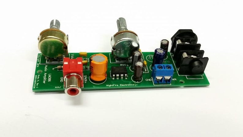 Puede incluir: Una placa de circuito verde con una toma RCA negra y roja, dos potenci&oacute;metros, dos condensadores, un bloque de terminales azul y una terminal de altavoz negra. La placa tiene el texto "Audio Amplifier LM386" y "Nightfire Electronics".