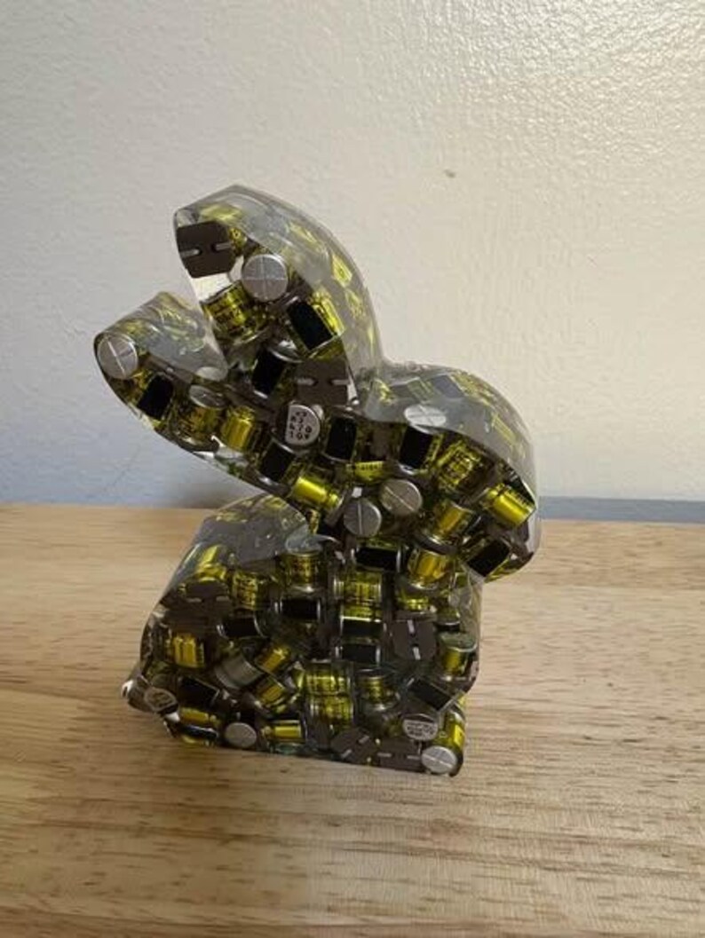 K&ouml;nnte beinhalten: Eine transparente Harzskulptur in Form eines Hasen, gef&uuml;llt mit kleinen, gelben, schwarzen und silbernen elektronischen Bauteilen. Die Skulptur steht auf einer Holzoberfl&auml;che. Einzigartiges Dekorationsst&uuml;ck.