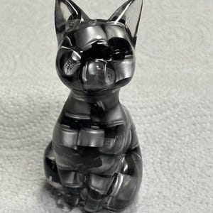 Puede incluir: Figura de gato de resina transparente en posici&oacute;n sentada. El gato est&aacute; relleno de componentes electr&oacute;nicos negros y plateados, creando un dise&ntilde;o abstracto. La figura tiene orejas puntiagudas y un acabado liso y brillante.