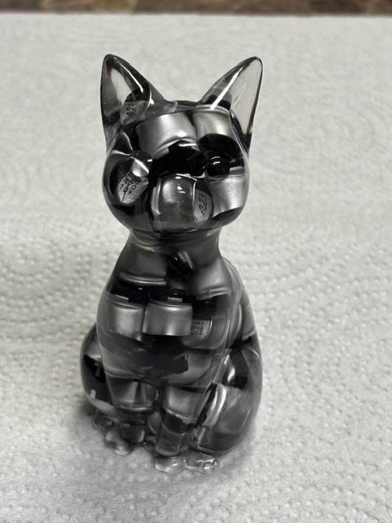 Puede incluir: Figura de gato de resina transparente en posici&oacute;n sentada. El gato est&aacute; relleno de componentes electr&oacute;nicos negros y plateados, creando un dise&ntilde;o abstracto. La figura tiene orejas puntiagudas y un acabado liso y brillante.