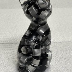 Puede incluir: Figura de gato de resina transparente con interior negro y plateado. El gato est&aacute; sentado con la cola enrollada. La figura est&aacute; hecha de resina transparente y llena de peque&ntilde;os componentes met&aacute;licos.