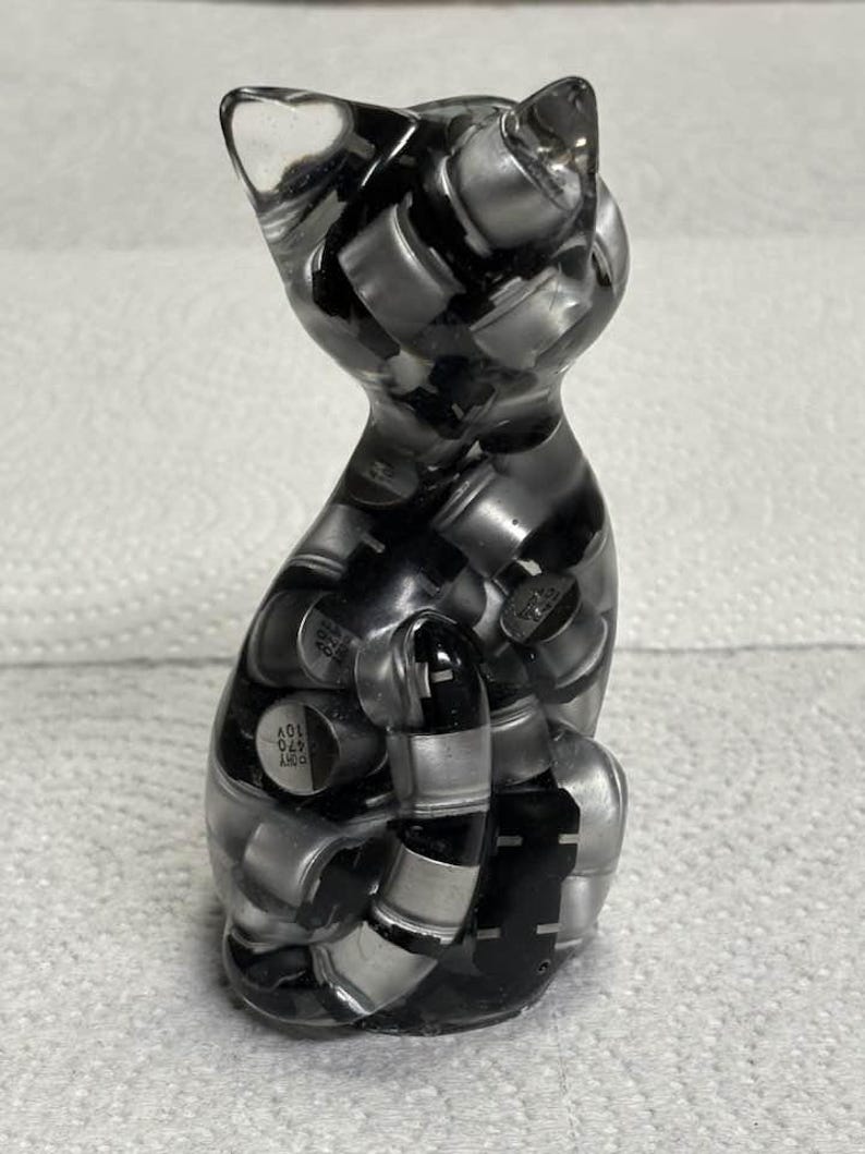 Puede incluir: Figura de gato de resina transparente con interior negro y plateado. El gato est&aacute; sentado con la cola enrollada. La figura est&aacute; hecha de resina transparente y llena de peque&ntilde;os componentes met&aacute;licos.