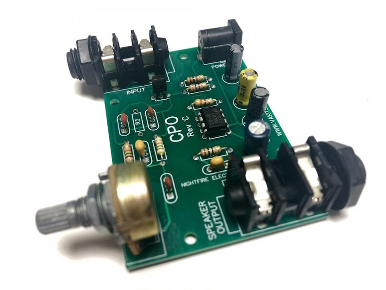 May include: A green circuit board with black and silver components. The board has a black knob, a black power jack, and two black audio jacks. The text "CPO Rev C" is printed on the board. The text "NIGHTFIRE ELEC" is printed on the board. The text "www.V-A-S-T.com" is printed on the board.