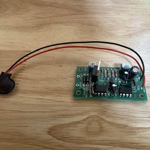 May include: A green electronic circuit board labeled American Siren with various components including resistors, capacitors, and integrated circuits, connected by red and black wires to a small black circular component on the left side.