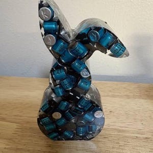 K&ouml;nnte beinhalten: Eine klare Harzskulptur in Form eines stilisierten Hasen. Die Skulptur ist mit zahlreichen kleinen, zylindrischen blauen und silbernen Komponenten gef&uuml;llt. Der Hase steht auf einer Holzoberfl&auml;che.