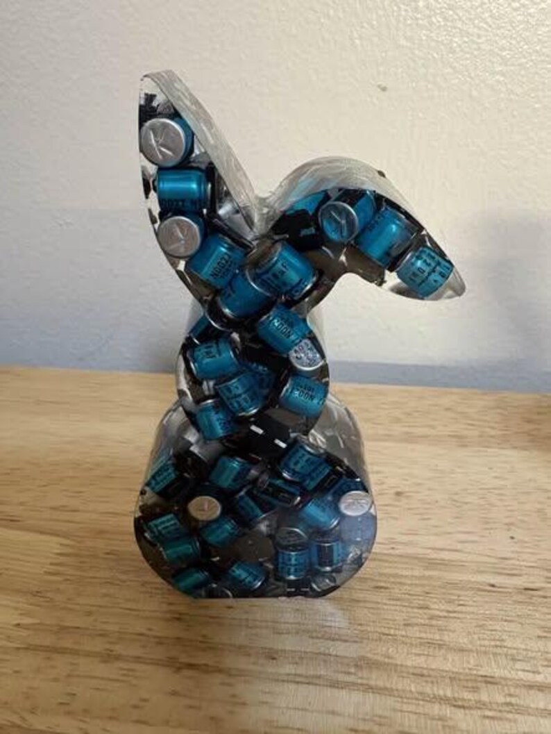 K&ouml;nnte beinhalten: Eine klare Harzskulptur in Form eines stilisierten Hasen. Die Skulptur ist mit zahlreichen kleinen, zylindrischen blauen und silbernen Komponenten gef&uuml;llt. Der Hase steht auf einer Holzoberfl&auml;che.